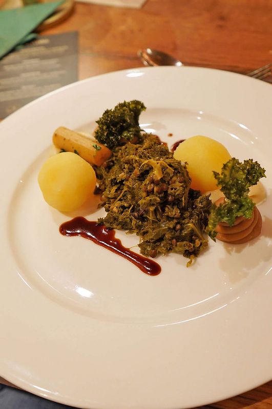 Teller mit Grünkohl, zwei Kartoffelklößen, einer Schwarzwurzel und einem Klecks dunkler Soße auf einem Holztisch.