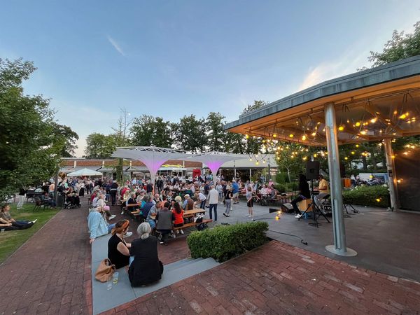 Menschen sitzen, stehen und tanzen beim Open-Air-Konzert am Zwischenahner Meer, Bühne mit Musiker und Lichterketten im Abendlicht.