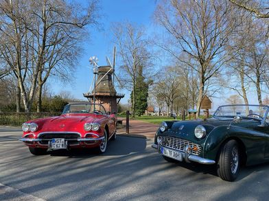 Roter Chevrolet Corvette und grüner Triumph Cabrio vor der Windmühle im Kurpark mit kahlen Bäumen.