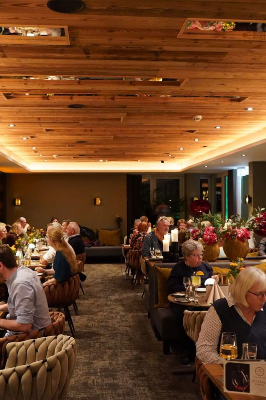 Innenraum des Restaurants Delikat in Bad Zwischenahn mit mehreren Gästen an gedeckten Tischen, warmes Holzdekor an der Decke und Blumenarrangements auf den Tischen.