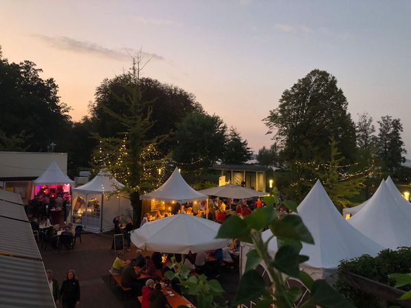 Weinfest am Meer bei Sonnenuntergang mit weißen Zelten, Lichterketten an Bäumen und Menschen an Tischen.