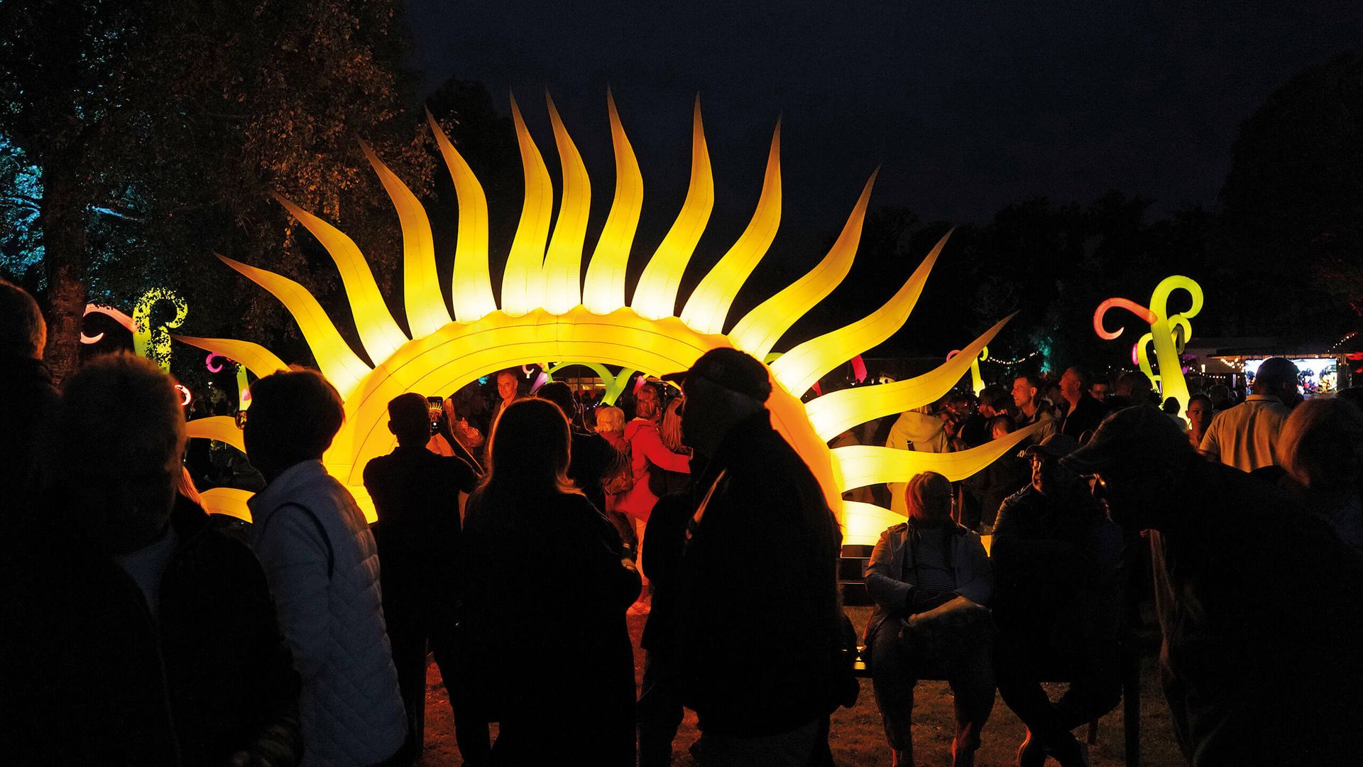 Leuchtende Sonnenskulptur mit strahlenförmigen Ausläufern vor dunklem Nachthimmel und Menschen im Vordergrund.