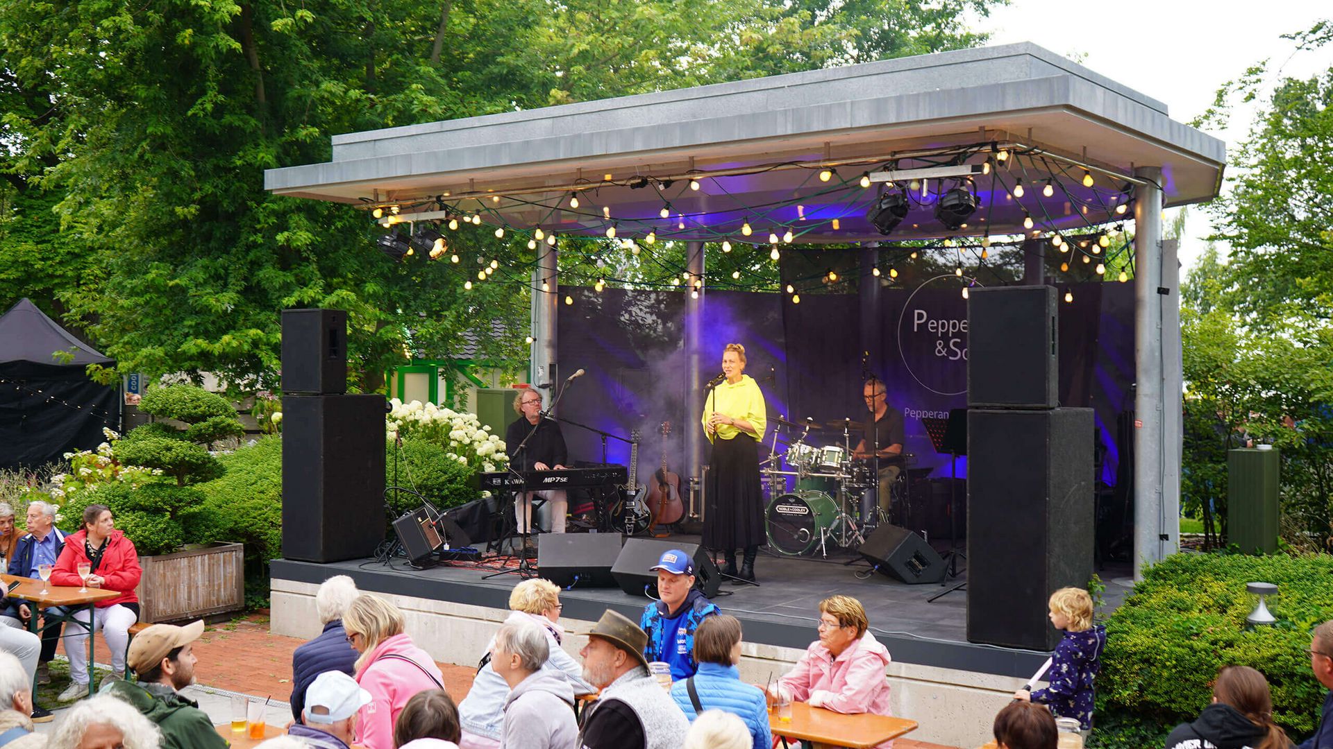 Freilichtbühne mit drei Musikern von Pepper & Soul, davor Publikum an Holzbänken auf der Terrasse der Wandelhalle, umgeben von Bäumen und Pflanzen.