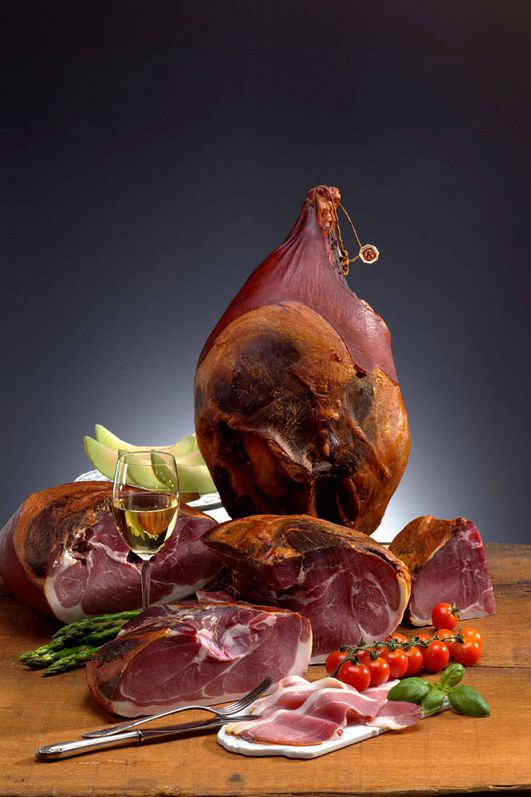 Verschiedene und unetrschiedlich große Schinkenstücke auf einem Tisch neben Tomaten, grünem Spargel und Wein.