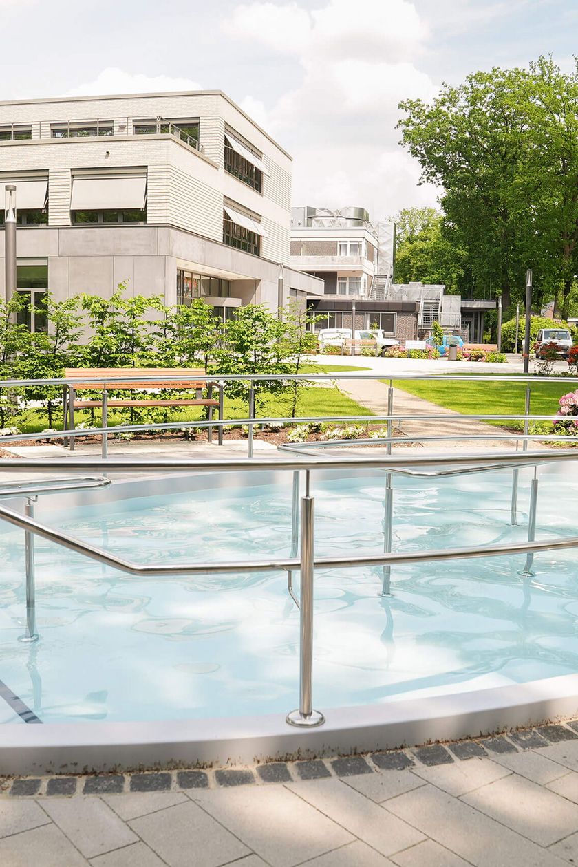 Kneippbecken im Sommer mit funkelndem Wasser und Sitzbank - im Hintergrund ein Klinik gebäude.