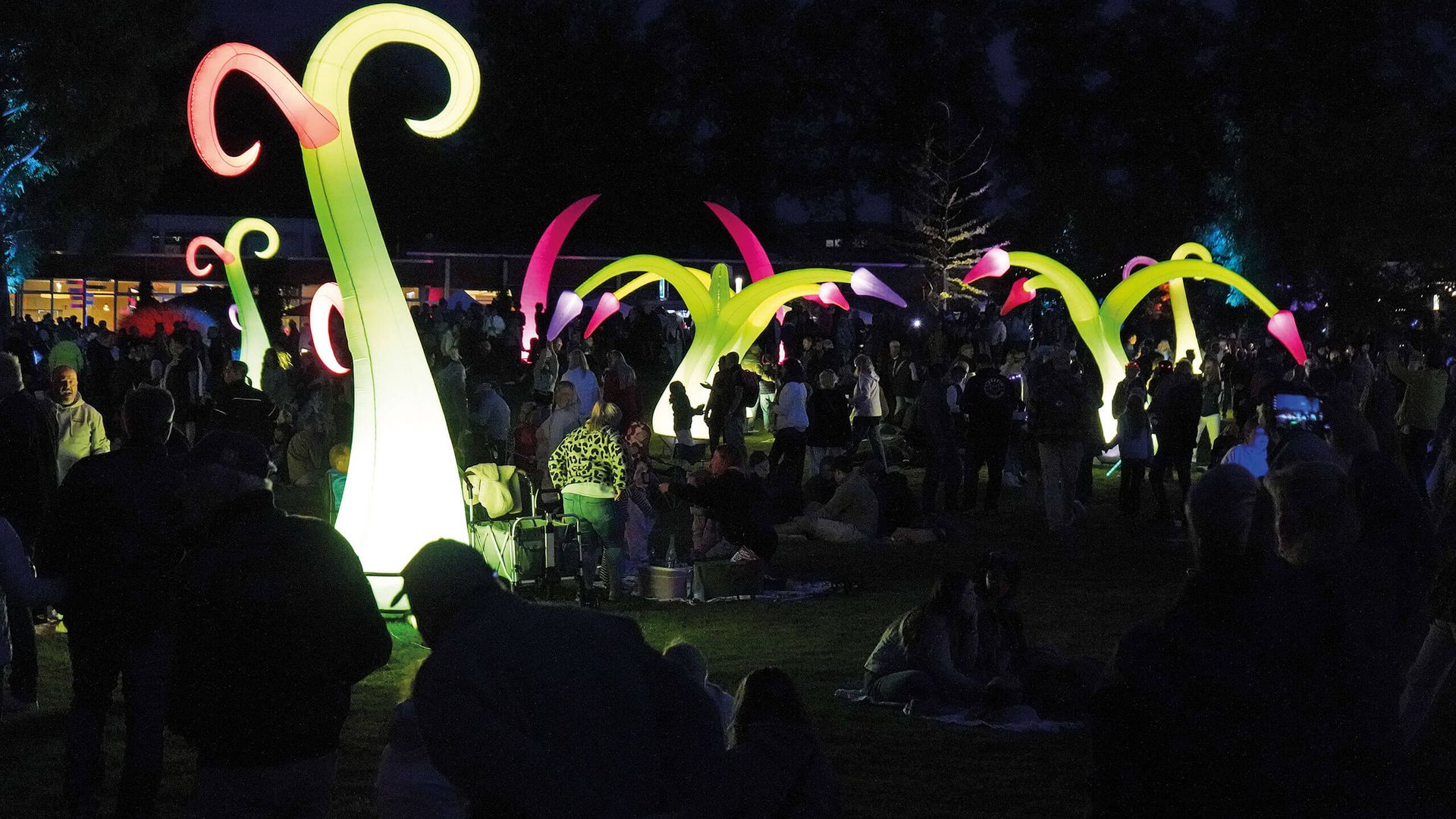 Leuchtende, gebogene Skulpturen in verschiedenen Farben im Kurpark vor einer Menschenmenge bei Nacht.