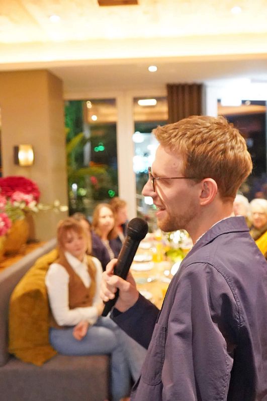 Dr. Grünkohl (Dr. Christoph Hahn) mit Mikrofon im Restaurant Delikat vor mehreren Gästen im Hintergrund.