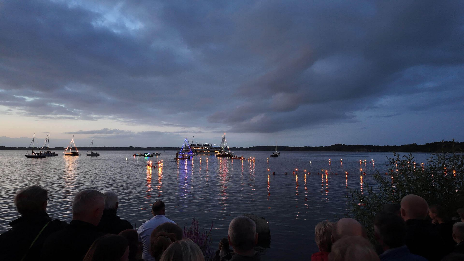 Menschen beobachten bei Dämmerung das Fackelschwimmen auf dem Zwischenahner Meer mit mehreren beleuchteten Booten und Fackeln im Wasser.
