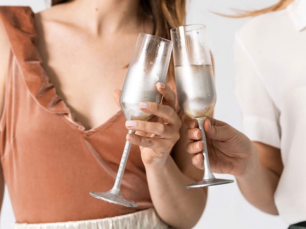 Zwei Frauen stoßen mit Sektgläsern an, eine trägt ein ärmelloses, rostfarbenes Oberteil mit Rüschen, die andere ein weißes Oberteil.