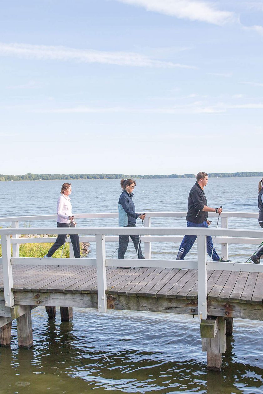 Mehrere Personen walken mit Stöckern über eine Brücke vor einem Schiff auf dem Zwischenahner Meer.