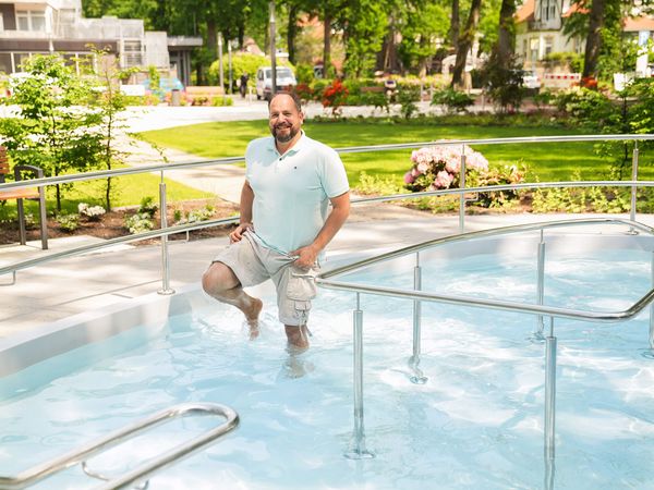 Ein Mann watet fröhlich in einem Kneippbecken, umgeben von blühender Natur und grünen Bäumen.