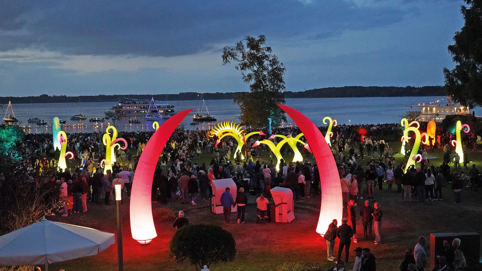Menschenmenge bei Dämmerung im Kurpark mit leuchtenden, bunten Lichtskulpturen und Wasser im Hintergrund.