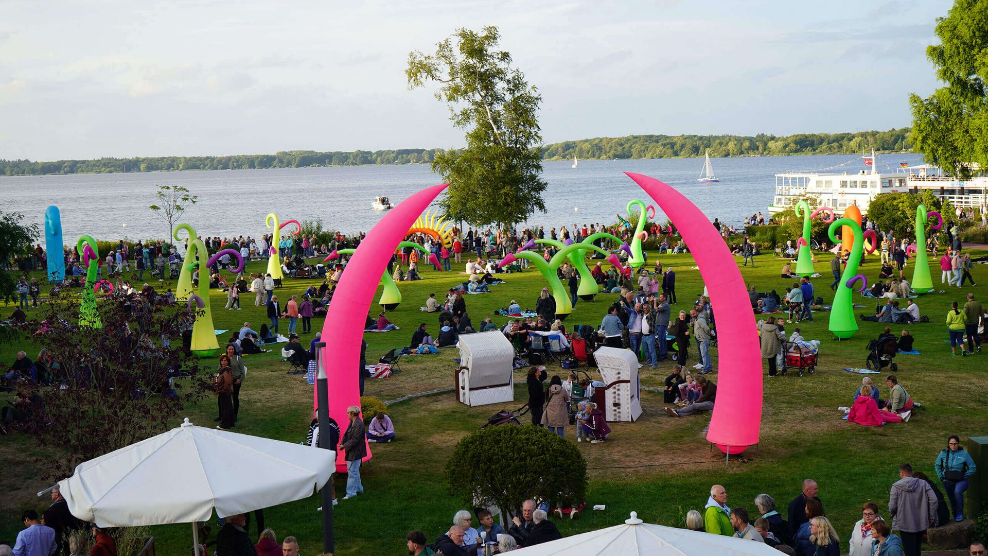 Menschen sitzen und stehen auf der Kurparkwiese am Seeufer zwischen großen, bunten, gebogenen Luftskulpturen bei Tageslicht.