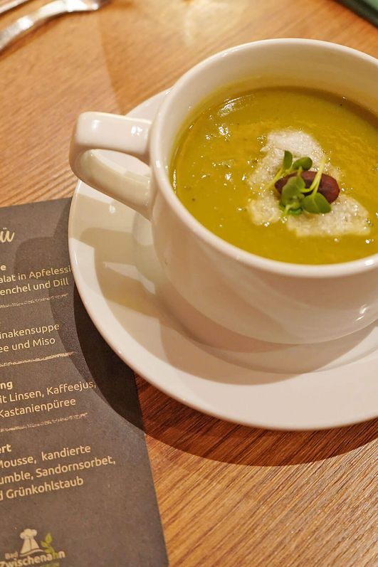 Suppentasse mit Grünkohlsuppe, garniert mit Sahne und Kräutern, auf weißem Unterteller auf Holztisch neben Besteck und Menükarte.