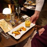 Hand eines Kellners serviert einen weißen Teller mit einem Glas und kleinen Portionen auf einem gedeckten Holztisch mit Weinglas, Besteck und Brotkorb.