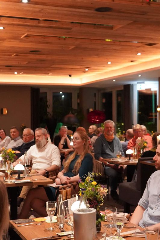 Frau hält ein Mikrofon und spricht vor Publikum in einem Restaurant mit Holzbalkendecke und gedeckten Tischen mit Blumen und Kerzen.
