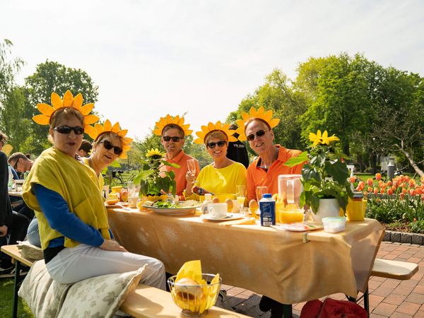 Fünf Personen mit Sonnenblumen-Kopfschmuck sitzen an einem gedeckten Tisch im Freien, umgeben von Blumen und grünen Bäumen, genießen ein Frühstück im Kurpark bei Sonnenschein.