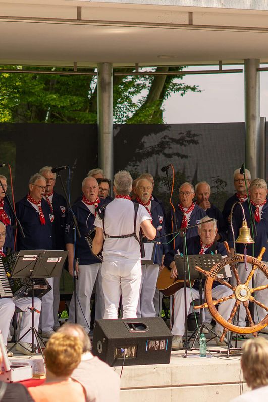 Ein Shanty-Chor tritt auf der Freilichtbühne im Kurpark auf, davor lauschen Zuschauer die Musik.