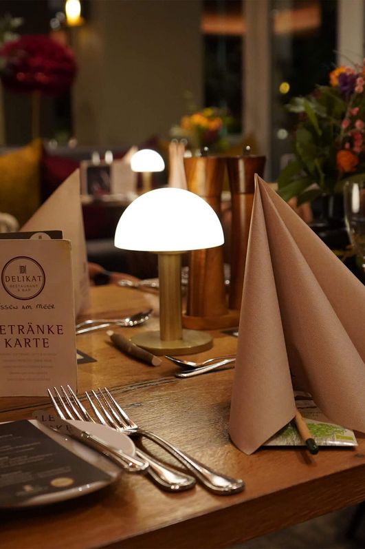 Tisch in einem Restaurant mit gefalteten Servietten, Besteck, Weingläsern, einer kleinen Lampe und einer Getränkekarte mit dem Schriftzug 'Getränke Karte'.