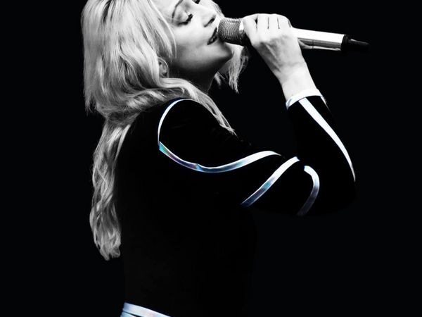 Die kleene Helene mit langen, welligen blonden Haaren hält ein Mikrofon nahe am Mund, trägt schwarzen Anzug mit silbernen Streifen auf Ärmeln und Taille.