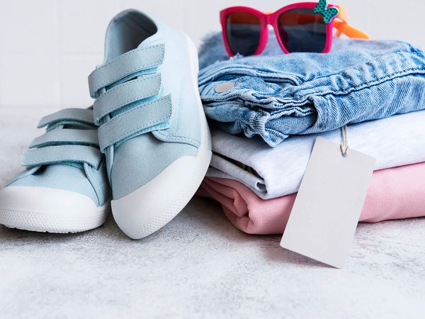 Hellblaue Kinderschuhe mit Klettverschluss neben gefalteter Kinderkleidung und einer pinken Sonnenbrille.