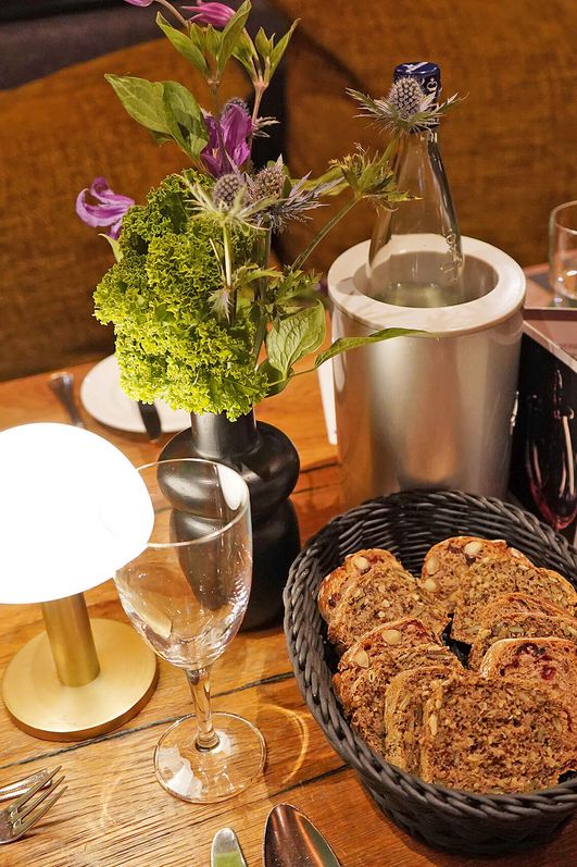 Gedeckter Holztisch mit grün gefalteten Servietten, Brotkorb, Gläsern, Besteck, einer kleinen Lampe und einem Blumenarrangement.