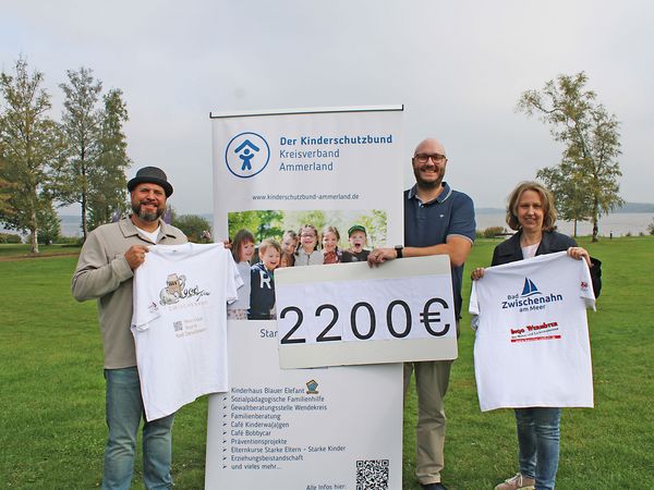 Drei Personen stehen auf einer Wiese vor einem Banner des Kinderschutzbundes Ammerland, halten ein Schild mit '2200€' und zwei weiße T-Shirts mit Logos und Texten zur Spendenübergabe.