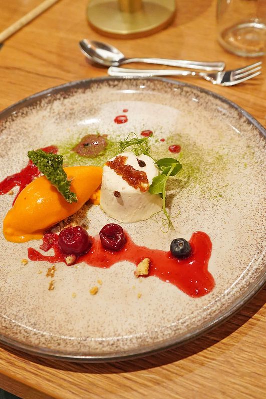 Dessert mit Vanille-Mousse, Sanddorn-Sorbet, roten Fruchtsoßen, Beeren und grüner Grünkohl-Dekoration auf einem runden Teller.