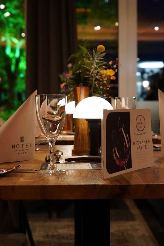 Gedeckter Holztisch mit zwei gefalteten weißen Servietten, Weingläsern, Besteck, Menükarte und kleiner Lampe im Restaurant Deilikat in Bad Zwischenahn.