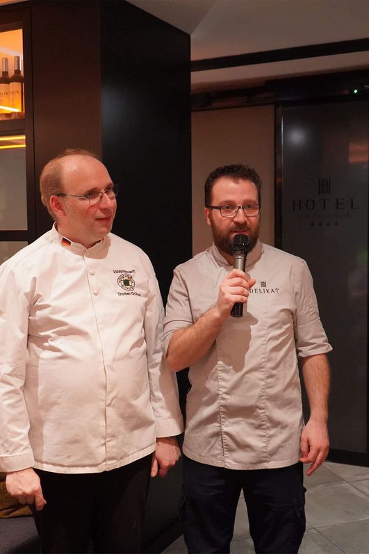 Zwei Männer in weißen Kochjacken, einer mit Mikrofon, stehen in einem Restaurant mit dunkler Einrichtung und beleuchtetem Regal im Hintergrund.