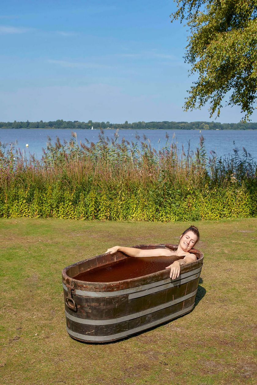 Eine Frau liegt in einer Holzwanne gefüllt mit Moor, die auf einer Wieser vor dem Ufer des Zwischenahner Meeres steht.