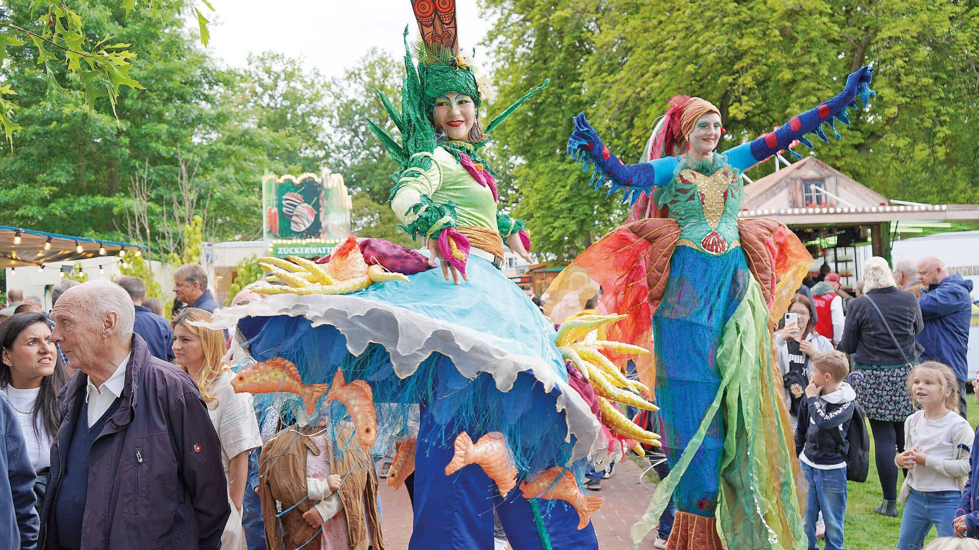 Zwei Stelzenläuferinnen in bunten fantasievollen Kostümen auf einem belebten Weg im Kurpark mit Zuschauern und Bäumen im Hintergrund.
