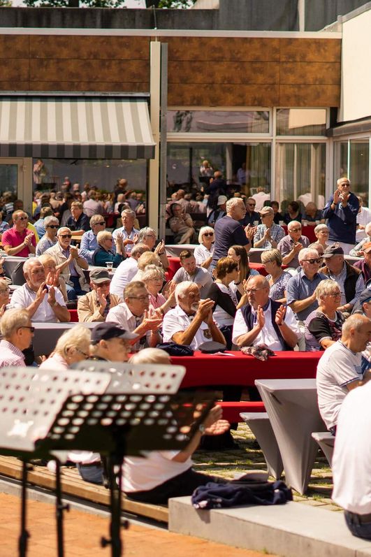 Große Gruppe von Menschen sitzt bei sonnigem Wetter auf Bänken und an Tischen im Freien vor der Wandelhalle und hört Musik von der Freilichtbühne im Kurpark.
