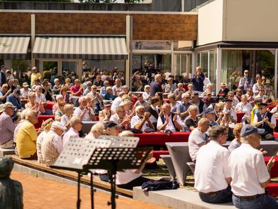 Große Gruppe von Menschen sitzt bei sonnigem Wetter auf Bänken und an Tischen im Freien vor der Wandelhalle und hört Musik von der Freilichtbühne im Kurpark.