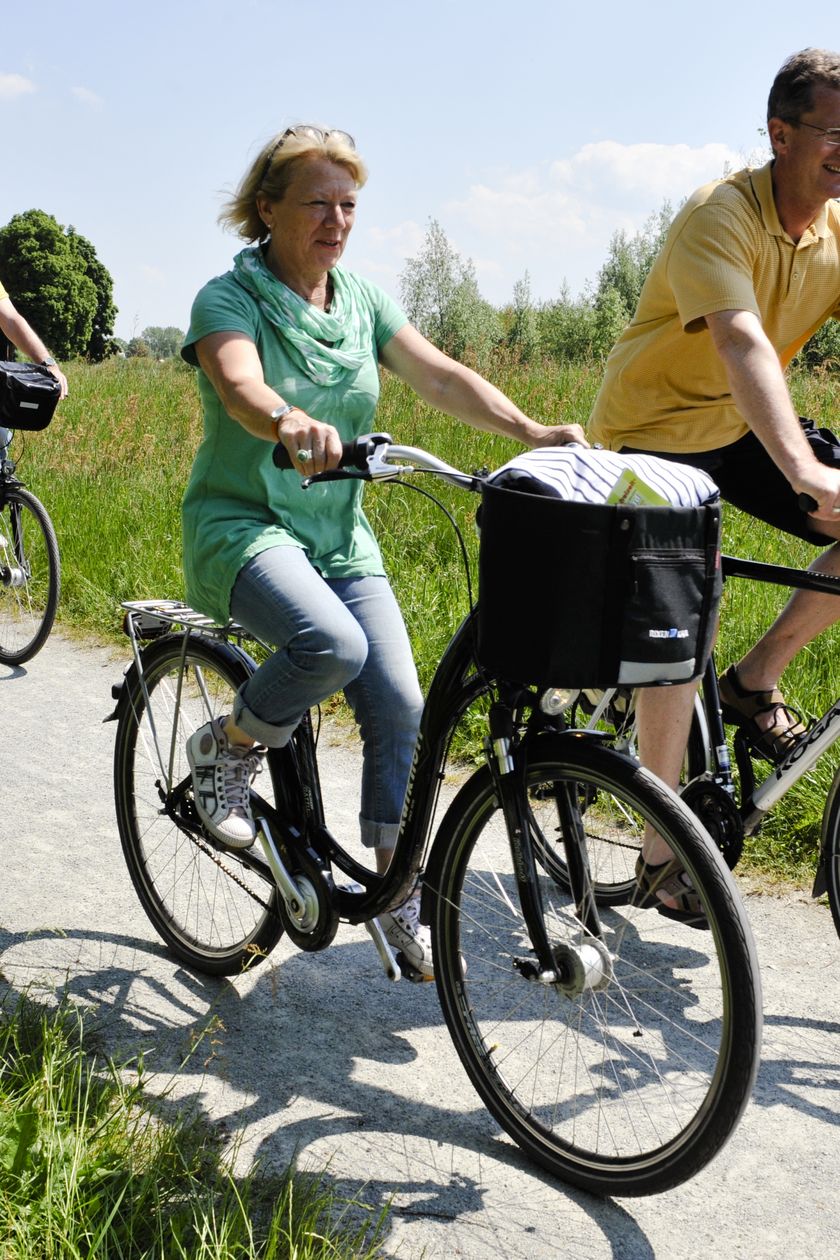Eine Gruppe aus vier Radfahrern fährt entlang eines Radweges bei sonnigem Wetter. Im Hintergrund steht eine schöne, alte Mühle