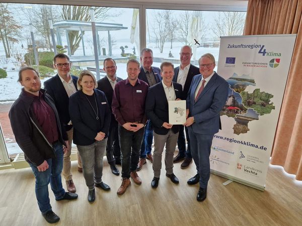 Gruppe von Personen in Innenraum vor Fenster mit Schnee draußen, eine Person hält eine Urkunde, daneben Roll-up mit Logo und Text 'Zukunftsregion 4 Klima'