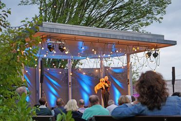 Blau beleuchtete Freilichtbühne beim Comedy-Open-Air auf dem der Bauchredner Marcelini mit einem Stoffhund steht, davor das Publikum sitzend auf Stühlen.